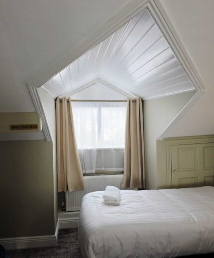 Cosy hotel room with soft white bedding, charming attic window, and complimentary towel, offering a serene stay.